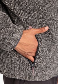Veste en polaire avec un extérieur gris texturé, présentant une main dans une poche latérale zippée et une doublure de poignet contrastante plus foncée.