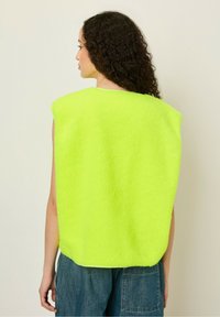 Neon yellow fleece vest with a boxy shape and no sleeves, textured fabric, and a round neckline. Worn over denim pants.