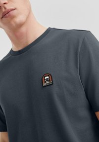 Grijze katoenen t-shirt met een ronde halsuitsnijding, voorzien van een klein bruin en crèmekleurig geborduurd logo op de borst. Glad textuur.