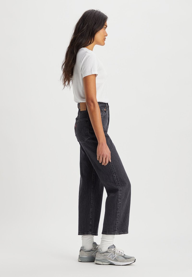 Jeans noirs taille haute avec une coupe droite, associés à un t-shirt blanc ajusté et des chaussures de sport grises. Design minimaliste.