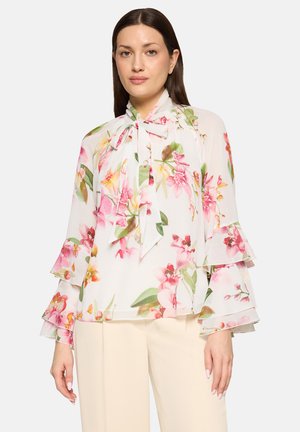 Kvinde iført en gennemsigtig hvid bluse med blomsterprint i lyserødt og grønt, lagdelte klokkeærmer og en bindebåndsudskæring, kombineret med cremefarvede bukser.