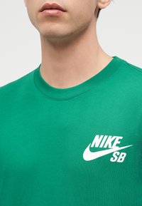Νέος που φοράει πράσινο t-shirt Nike SB με στρογγυλή λαιμόκοψη, με το λογότυπο της μάρκας στο αριστερό στήθος.