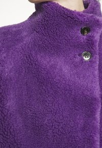 Lila fleecejacka med hög krage, med en texturerad yta, två svarta knappar och en rak silhuett.