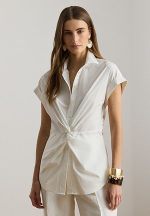 Kvinde iført en hvid kortærmet skjorte med knapper, snoet ved taljen, hvide bukser, guld- og skildpaddemønstrede armbånd samt store øreringe af muslingeskaller.