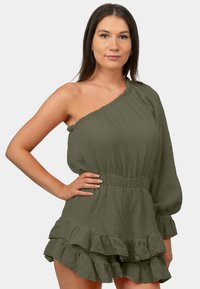 Combinaison une épaule verte en tissu texturé, avec un ourlet volanté et une taille élastique. Présente un design fluide avec des manches longues.