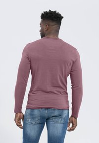 Langärmliges Shirt in staubrosa Farbe, aus weichem Stoff. Rundhalsausschnitt und taillierte Silhouette. Getragen mit hellblauen Jeans.