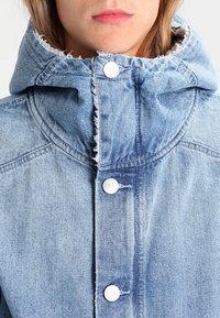Light blue denim jacket featuring a hood, five silver snap buttons, and frayed edges along the collar and hem.