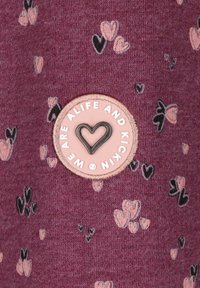 Tessuto bordeaux con motivi a cuore sparsi in rosa e nero. Presenta una toppa rotonda, rosa chiaro, con testo nero e un'accento a forma di cuore.