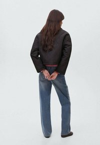 Jachetă neagră matlasată, scurtă, cu mâneci late, asortată cu blugi bleu deschis, cu talie înaltă. Modelul are părul lung și mâinile încrucișate în spate.