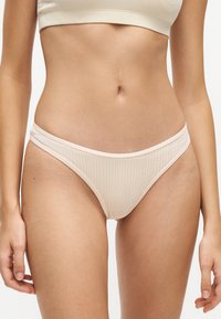 Torse féminin portant un bas de bikini côtelé beige clair et un haut assorti sur un fond blanc uni.