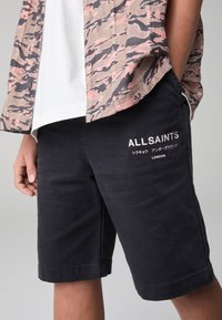 Čierne bavlnené šortky s potlačou loga a textu na jednej strane. Na biele tričko je nosená ľahko vzorovaná košeľa rozopnutá.