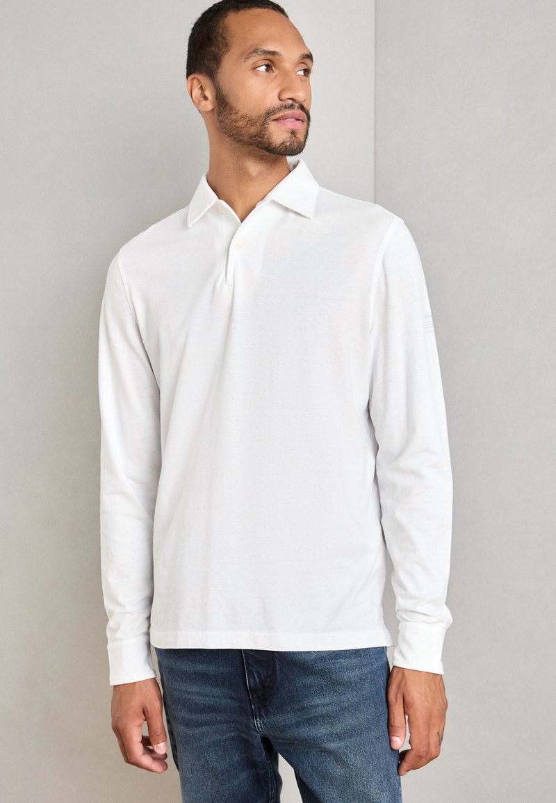 Mann, der ein weißes Langarm-Polohemd und blaue Jeans trägt, steht vor einer schlichten hellgrauen Wand und schaut zur Seite.