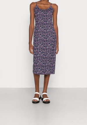 Femme portant une robe mi-longue sans manches bleu marine avec un petit imprimé floral rose et blanc, associée à des sandales blanches à bout ouvert.