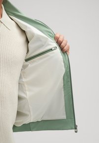 Interno di giacca verde chiaro con un tessuto liscio. Presenta una sezione foderata di bianco con una tasca con zip. Design minimalista e cuciture pulite.