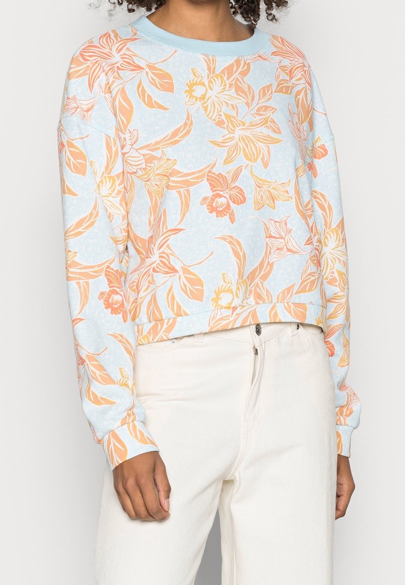 Sudadera azul claro con patrones naranjas y florales, que presenta un cuello redondo, mangas largas y un dobladillo cortado. Combinada con pantalones blancos.