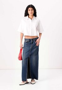 Camisa branca curta, saia de denim de cintura alta com racha frontal, ajuste moderno. Modelo segura uma bolsa vermelha, usando sapatos claros.