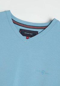Jasnoniebieska bawełniana koszulka V-neck o gładkiej teksturze. Posiada subtelne logo oraz paskowany akcent na wewnętrznym kołnierzyku. Rozmiar L.