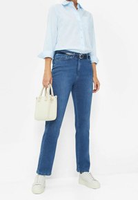 Chemise bleu clair à manches longues, jean bleu foncé coupe droite, tenant un petit sac à main blanc, et baskets blanches, sur un fond uni.