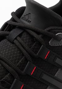 Chaussure de sport noire avec une tige en maille, surface texturée, lacets noirs et accents rouges. Présente un col rembourré et un détail de logo de marque.