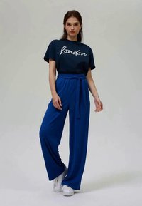 Μπλουζάκι ναυτικού στιλ με εκτύπωση "Λονδίνο", συνδυασμένο με φαρδιά μπλε παντελόνια με κορδόνι στη μέση. Το μοντέλο φοράει λευκά αθλητικά παπούτσια.