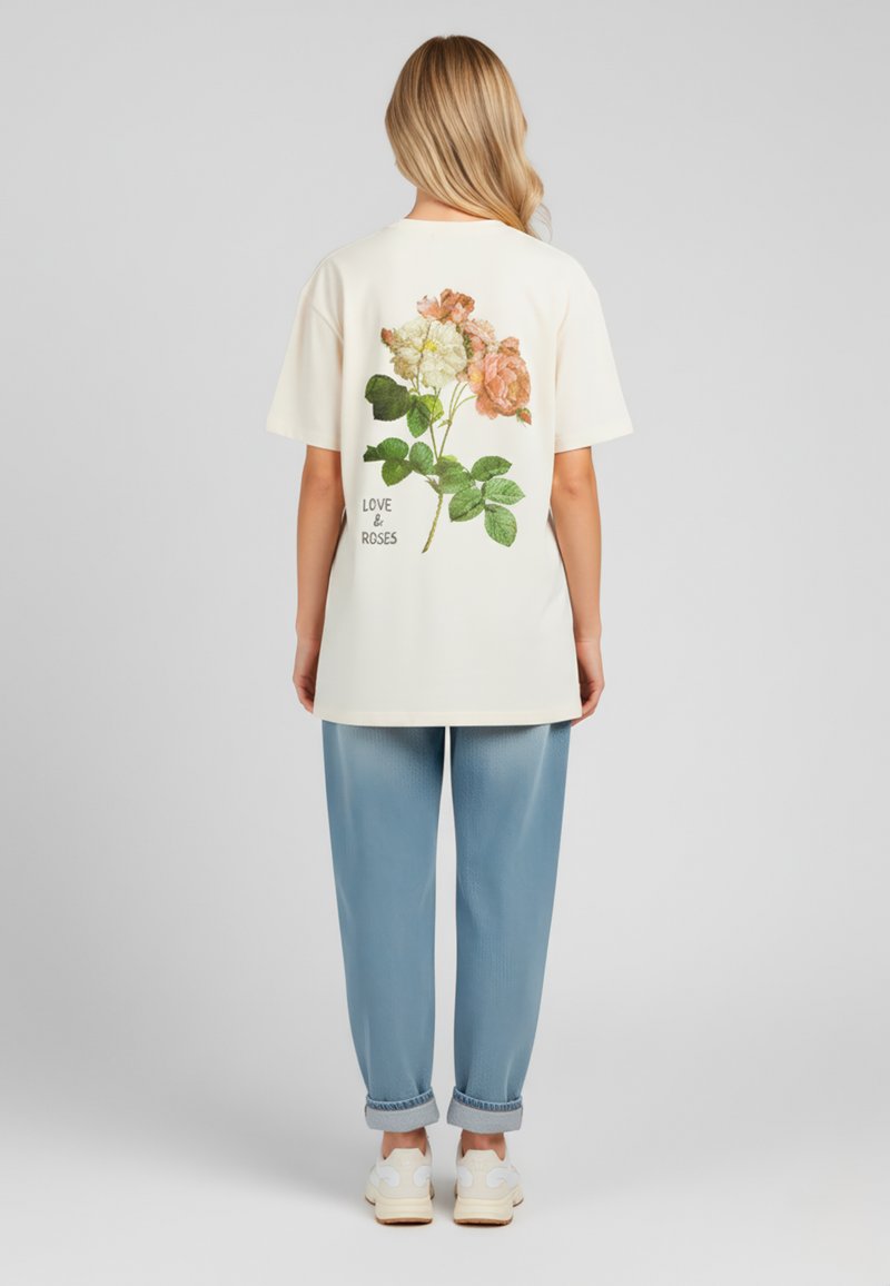 Camiseta de crema claro con un estampado floral de rosas rosa y blancas y hojas verdes en la parte de atrás, presenta el texto "AMOR Y ROSAS". Combinada con pantalones azules.