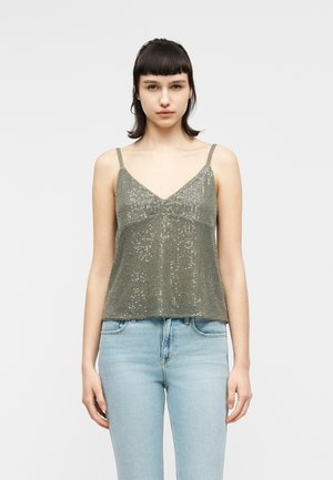 Mladá žena s krátkymi tmavými vlasmi, oblečená v zelenom flitrovanom topu na tenké ramienka a svetlomodrých džínsach, stojaca pred jednoduchým pozadím.