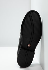 Suola di scarpa in gomma nera con motivi di presa testurizzati e un dettaglio con logo circolare. Sono visibili bordi lisci e un design della punta arrotondata.