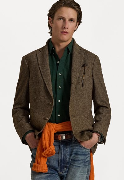 Blazer en tweed marron, chemise à boutons verte, pull orange noué à la taille et jean bleu. Comprend des poches et une montre au poignet.