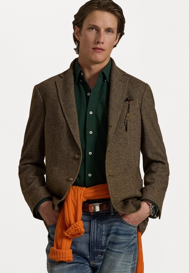 Brun tweed-blazer, grønn skjorte med knapper, oransje genser bundet rundt livet, og blå jeans. Funksjoner inkluderer lommer og klokke på håndleddet.