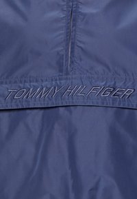 Granatowa kurtka wiatrówka o gładkiej, błyszczącej fakturze z haftem "TOMMY HILFIGER" w kontrastującym kolorze wzdłuż dołu.