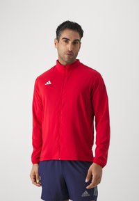 Czerwona sportowa kurtka z wysokim kołnierzem, zamkiem błyskawicznym i elastycznymi mankietami. Posiada biały emblem z trzema paskami po lewej stronie klatki piersiowej. Wykonana z lekkiego materiału.