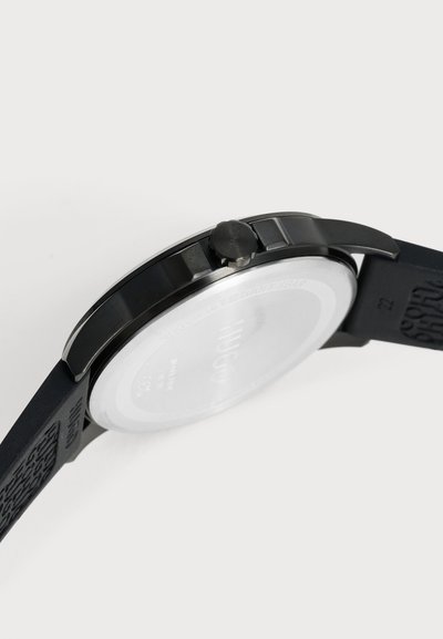 Gros plan sur une montre-bracelet noire, montrant son côté, sa couronne, le fond du boîtier et son bracelet en caoutchouc texturé sur un fond clair.