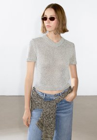 Maglione corto a maniche corte in argento con scollo a V, abbinato a jeans azzurri a vita alta e una cintura in maglia intrecciata in vita.