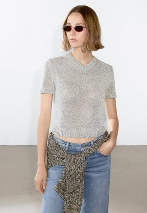 Silver short-sleeve cropped knit sweater with a V-neck, paired with high-waisted light blue jeans and a textured knit wrap at the waist.