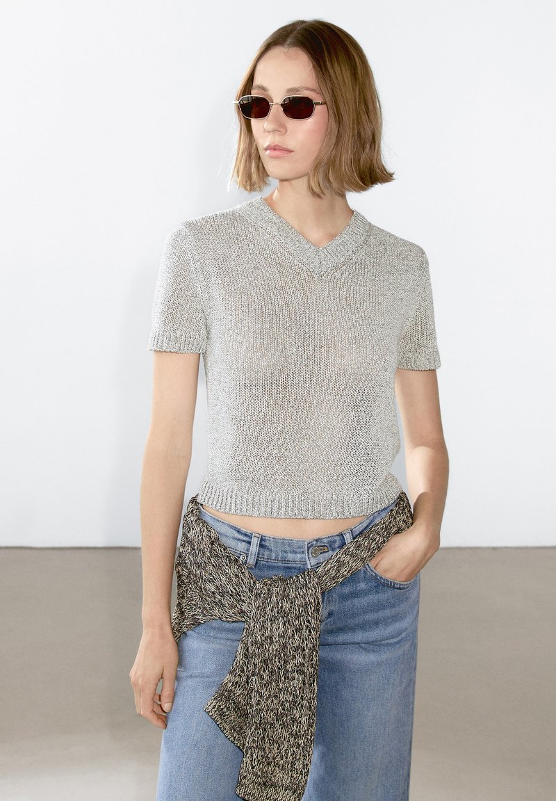 Maglione corto a maniche corte in argento con scollo a V, abbinato a jeans azzurri a vita alta e una cintura in maglia intrecciata in vita.