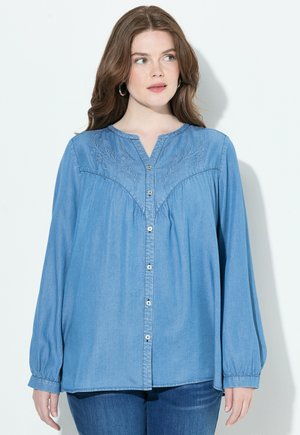 Vrouw in een lichtblauwe blouse met lange mouwen en knopen, versierd met bloemenborduursel op de borst, in combinatie met donkerblauwe jeans.