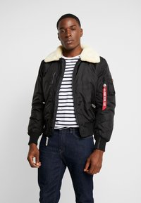 Chaqueta bomber negra con un cuello de borreguito crema, bolsillos con cremallera y una etiqueta de accesorio roja; llevada sobre una camisa a rayas azul marino y blanca.