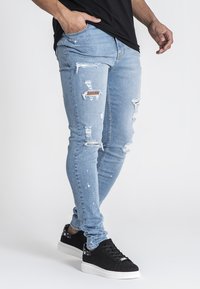 Smal passform ljusblå jeans med slitna hål, i kombination med mörka fläckar och färgstänk, tillsammans med svarta sneakers.