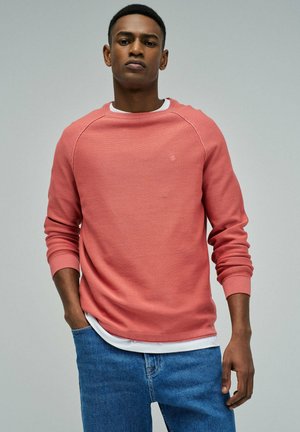 Joven con suéter de manga larga color salmón sobre una camisa blanca y jeans azules, de pie con una mano en el bolsillo sobre un fondo liso.