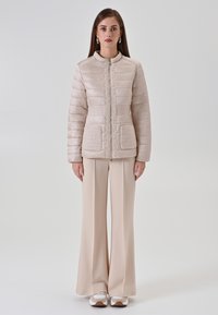 Doudoune beige avec coutures horizontales, accents de poche texturés, col montant et fermeture éclair avant. Associée à un pantalon beige à jambes larges.