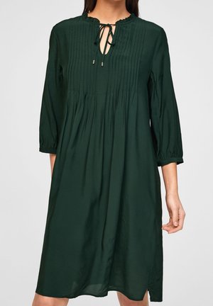 Robe verte longueur genou avec manches trois-quarts, plis sur le devant et col en trou de serrure noué sur une personne debout.