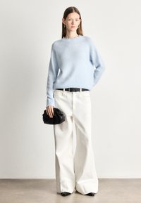 Světle modrý pletený svetr s kulatým výstřihem, doplněný širokými bílými kalhotami. Modelka drží malou černou tašku.