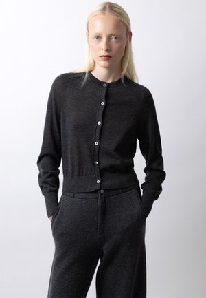 Blonde Frau mit blauen Augen, die eine dunkelgraue, geknöpfte Strickjacke und passende Hose trägt, stehend mit den Händen in den Taschen vor weißem Hintergrund.