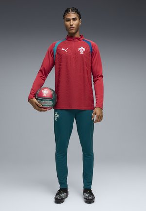 Joven atleta masculino con uniforme de fútbol de Portugal en rojo y verde, sosteniendo un balón de fútbol a juego sobre un fondo gris.