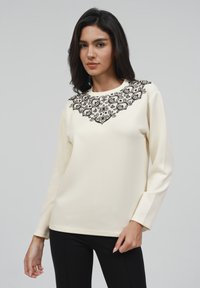 Crème lange mouw top met een zwart geborduurd bloemenontwerp rond de halslijn en een soepele, lichte stof met een slim fit.