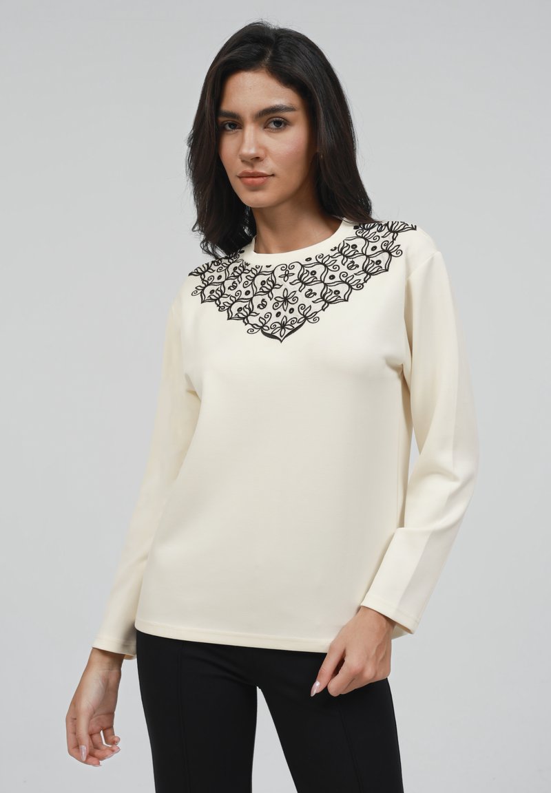 Crème lange mouw top met een zwart geborduurd bloemenontwerp rond de halslijn en een soepele, lichte stof met een slim fit.