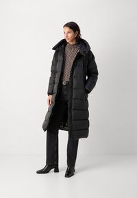 Langer schwarzer Puffer-Mantel mit Kapuze, mit einem gesteppten Design und einem Reißverschluss vorne. Getragen über einem schwarz-weiß gestreiften Rollkragenpullover und schwarzen Jeans.