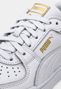 Vit sneakersko med guldfärgat Puma-logotyp, lädermaterial, perforerad tåbox, platta skosnören och texturerade detaljer. Klassisk lågprofilmodell.