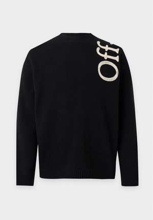 Jeune homme debout portant un pull noir avec le texte "Off" et un pantalon de survêtement gris clair avec un logo bleu, ainsi que des baskets blanches sur un fond uni.