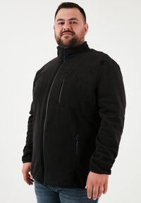 Chaqueta de forro polar negra con un cuello alto, bolsillos con cremallera y cierre frontal con cremallera. Textura suave, diseño casual y ajuste ajustable.